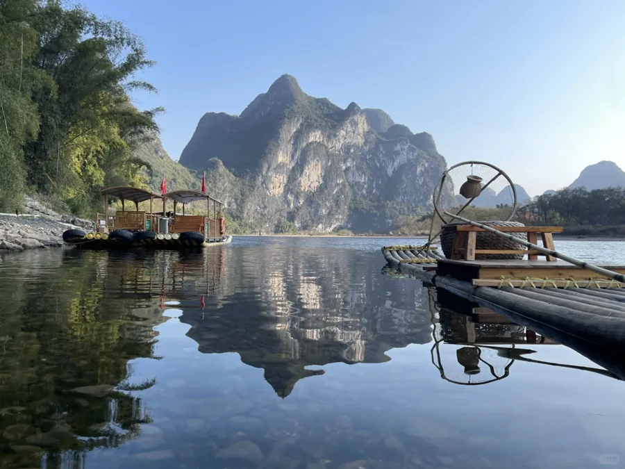 Still river reflecting karst hills, moored covered boats and a bamboo raft on a clear day